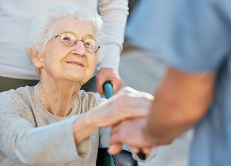 Holding hands, caregiver and senior woman in wheelchair for support outdoor in retirement home. Lov