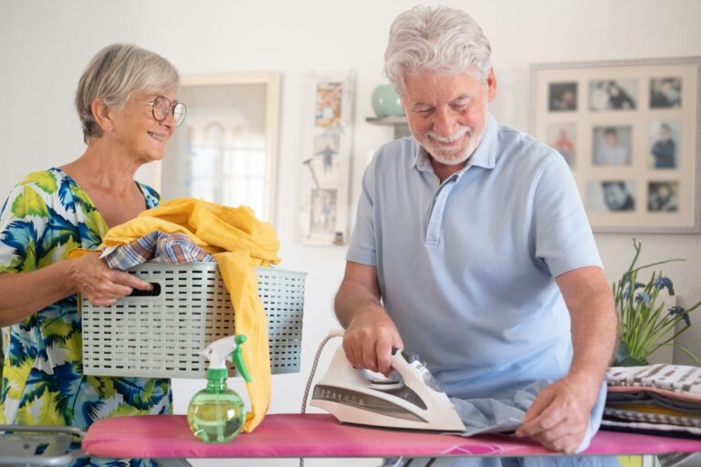 Smiling elderly man helps with housework by ironing clothes on the ironing board